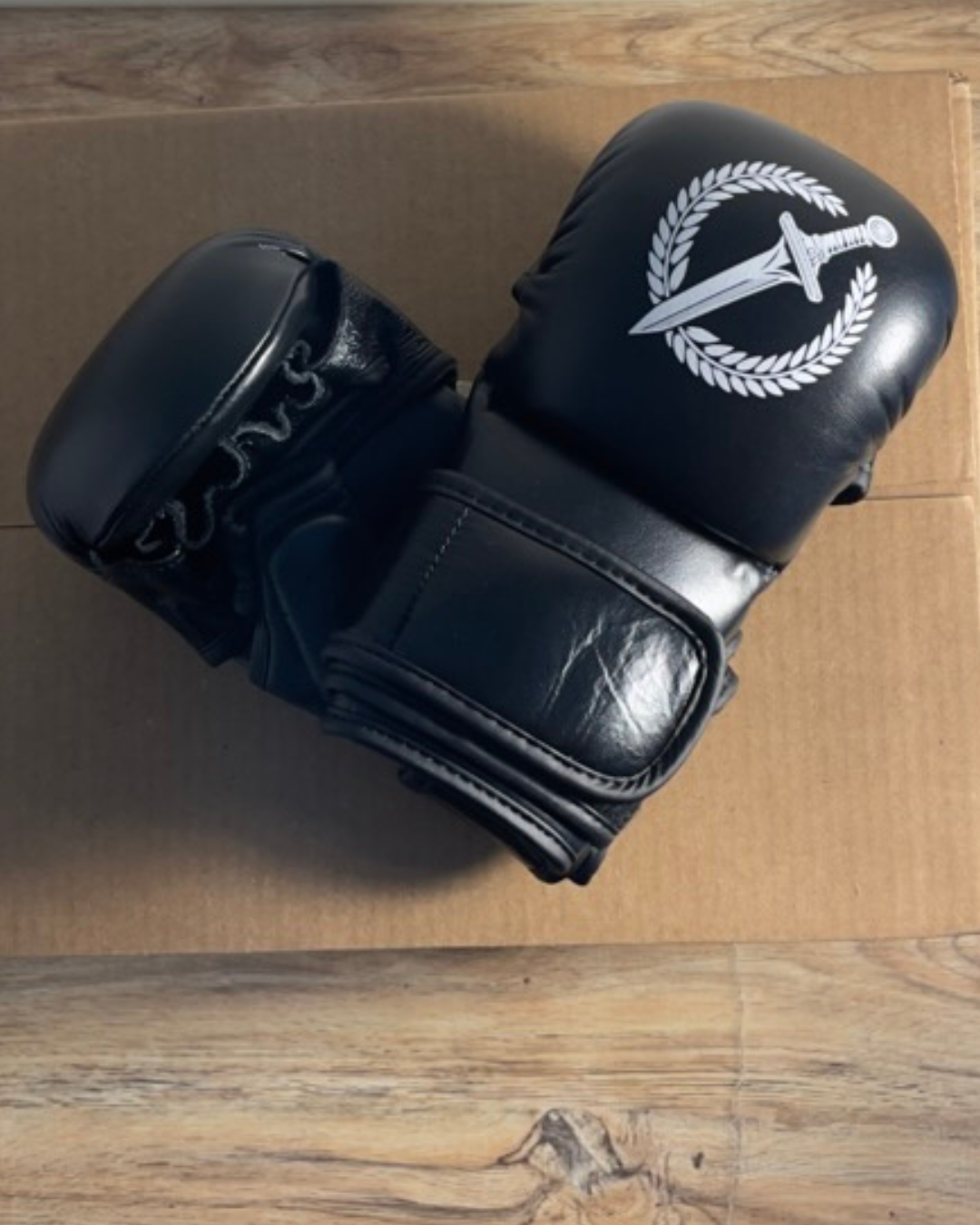 Black MMA gloves with a logo on top of a cardboard box on a wooden floor.