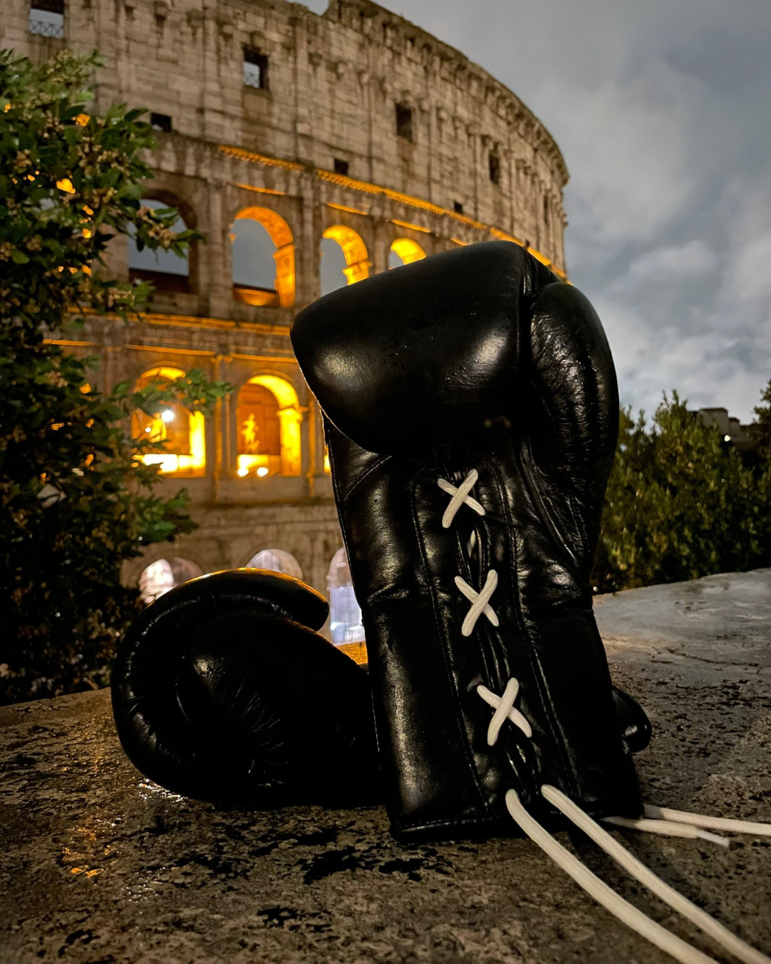 Black boxing gloves with white laces in front of the Colosseum at night.
