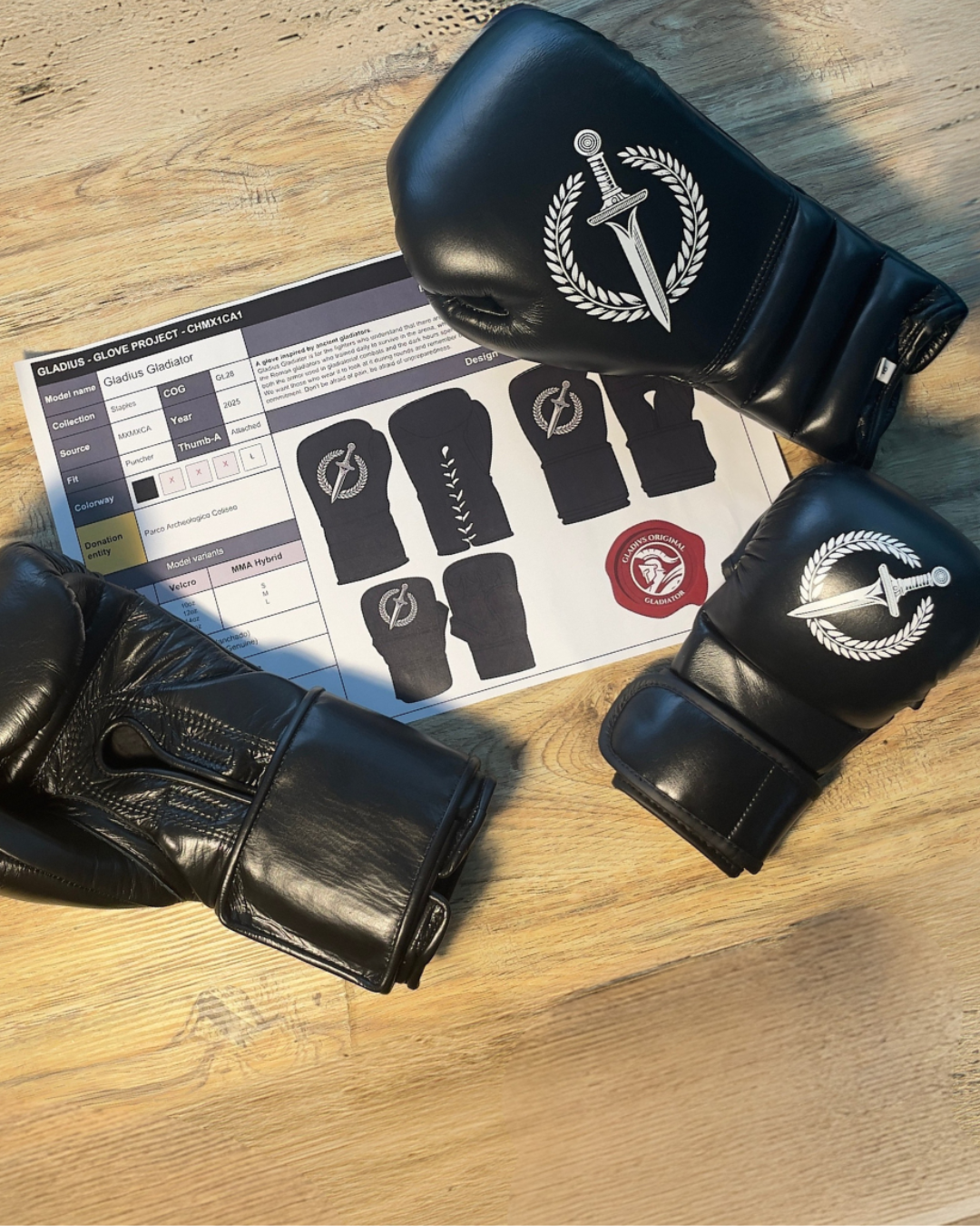 Black boxing gloves with a logo on a wooden surface next to a product sheet.