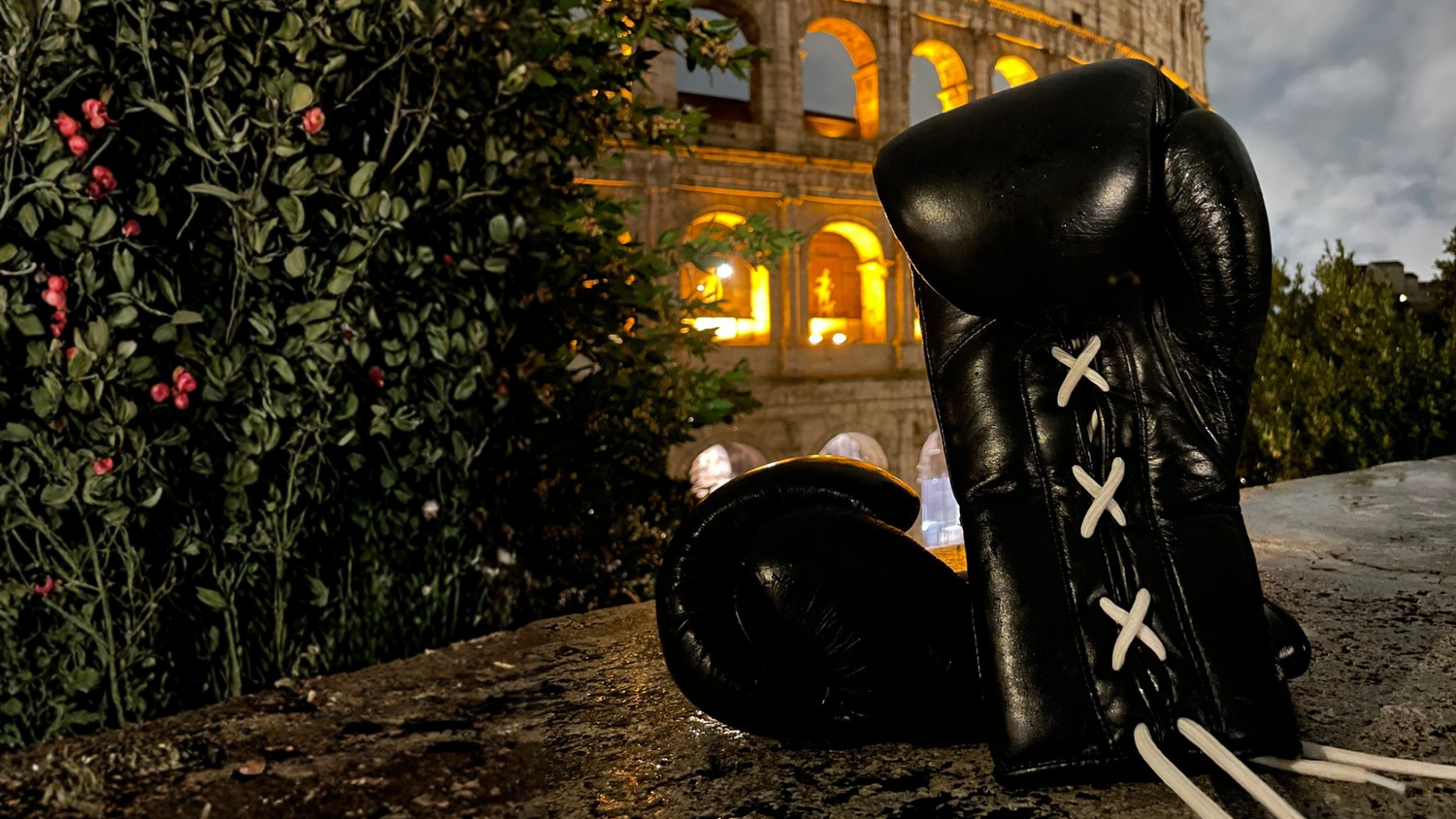 Black boxing gloves with white laces on a stone ledge in front of a historic building at night.